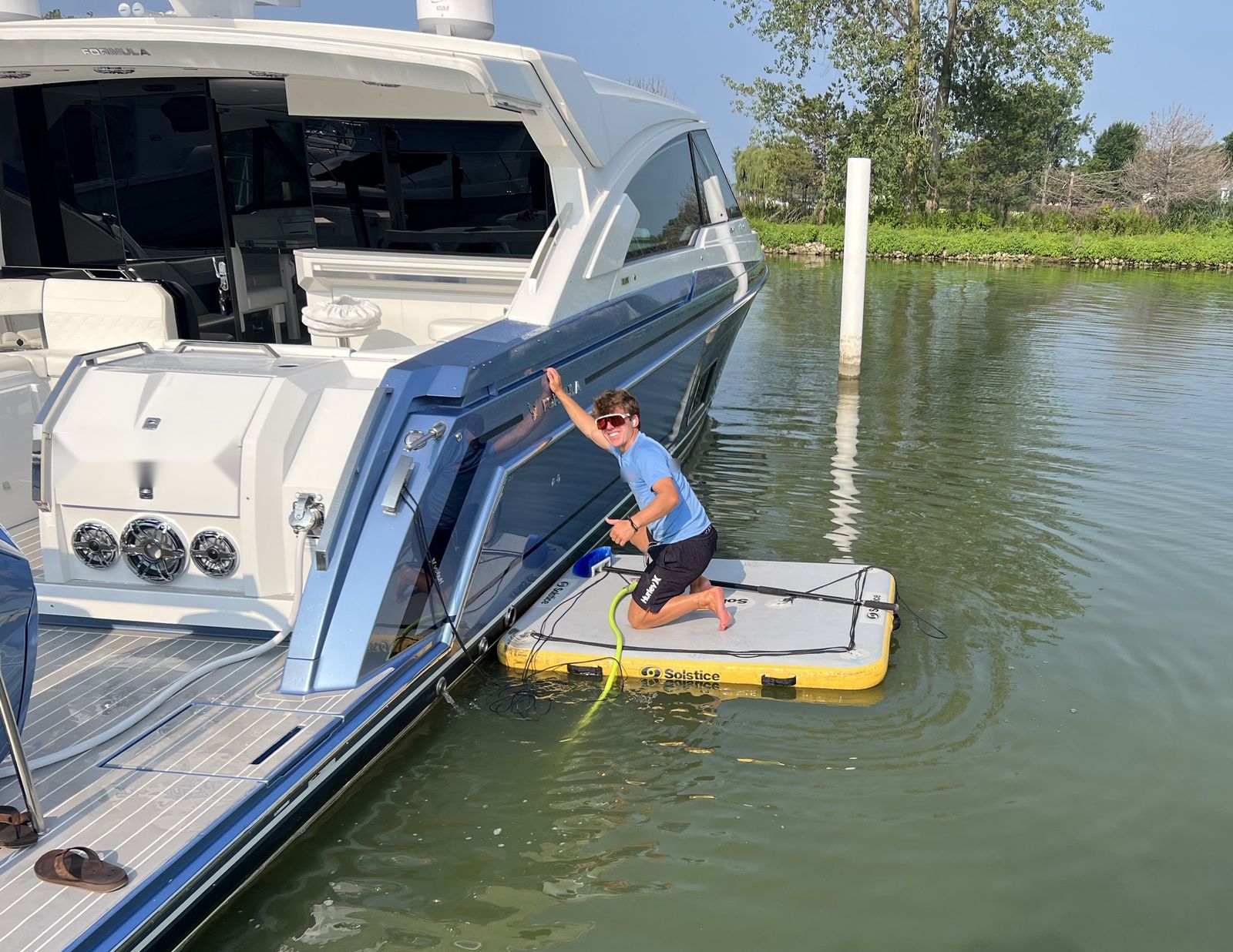 A detailer kneeling on an inflatable work dock beside a cruiser's hull, with a soft brush and a hose